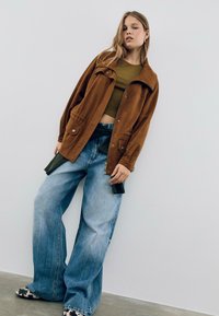 Veste en daim marron avec poches, associée à un jean bleu clair taille haute et ample. Modèle portant un crop top vert et des chaussures à motifs.