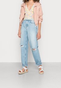 Lyserød zip-up jakke over en pastelfarvet blomsterbluse, parret med lyseblå ripped jeans og hvide sandaler med jute-sål.