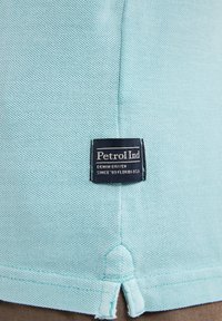Polo shirt azzurro chiaro con tessuto texturizzato, dotato di un'etichetta nera con la scritta "Petrol Ind" e dettagli di cucitura vicino all'orlo.
