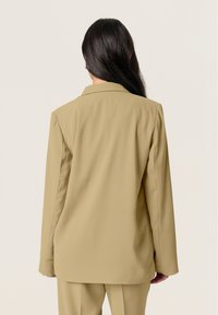 Beige blazer with a notched collar, made of smooth fabric. Features straight cut and slightly elongated sleeves, displaying a minimalist design.