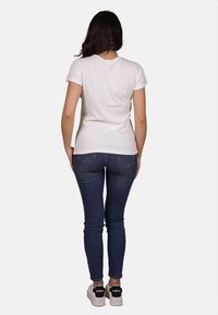 T-shirt bianco a maniche corte abbinato a jeans skinny blu scuro e sneakers nere. Modello visto da dietro. Texture del tessuto liscia.