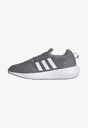 Šedý Adidas teniskový model s bílou podrážkou a třemi bílými pruhy na pleteném svršku, pohled z boku na bílém pozadí.