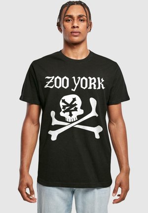 Musta puuvillapaita, jossa on valkoinen graafinen kuvitus pääkallosta ja ristikäärmeistä, ja tekstinä "ZOO YORK" yläpuolella. Lyhyet hihat, rento istuvuus, pyöreä pääntie.