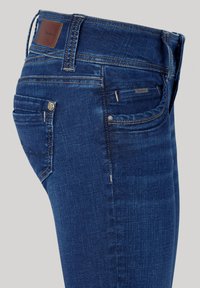 Dark blue denim jeans with a smooth texture, featuring a leather logo patch on the back waist and subtle stitching details.