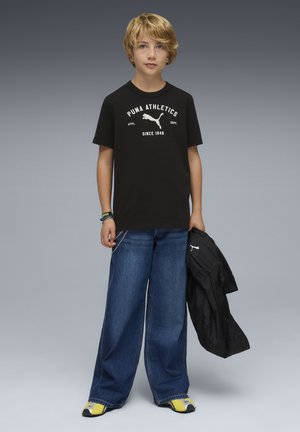 Camiseta negra con gráfico blanco "PUMA ATHLETICS", combinada con vaqueros azules sueltos y zapatos negros con acentos amarillos, sosteniendo una chaqueta negra.