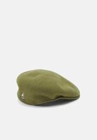 Beret en laine verte avec une texture lisse, forme ronde et une petite patte de logo blanche sur le côté. Design simple et discret.