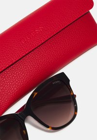 Ochelari de soare negri și țestoasă cu lentile gradient, alături de un toc texturat din piele roșie, embosat cu "GUESS."
