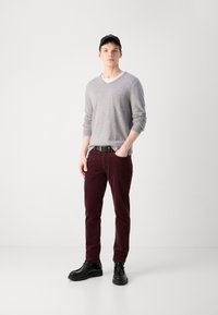 Pull en V gris, pantalon en velours côtelé bordeaux et chaussures en cuir noir. Le modèle porte une casquette noire ; style décontracté, design simple et silhouette ajustée.