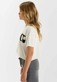 T-shirt blanc court avec logo noir brodé sur le devant, associé à un jean gris taille haute présentant une poche arrière et un détail de nœud.