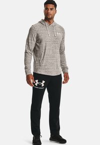 Sudadera gris con capucha y cordones, combinada con pantalones deportivos negros que cuentan con un logo blanco en el muslo izquierdo. Tejido ligero y deportivo.