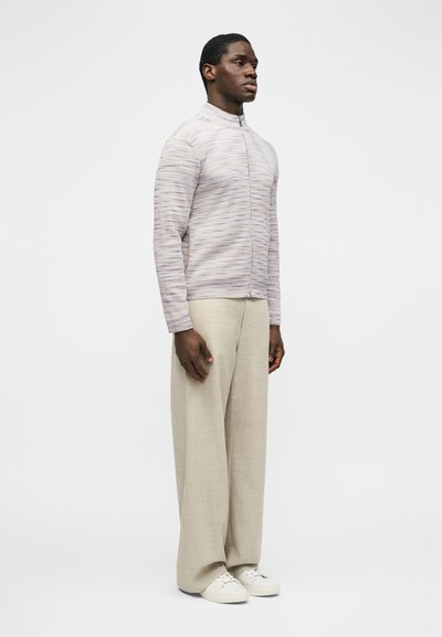 Homme debout, portant un pull zippé gris clair, un pantalon beige à jambes larges et des baskets blanches, sur un fond blanc uni.
