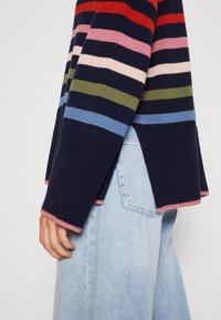 Randig tröja i marinblått, rosa, rött, grönt och blått med horisontella ränder; ribbade muddarna har rosa detaljer; lagd över ljusa jeans.