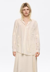 Camicia button-up beige chiaro con ricami floreali sulle maniche, una tasca frangiata e tessuto morbido e textured. Abbinata a una gonna coordinata.