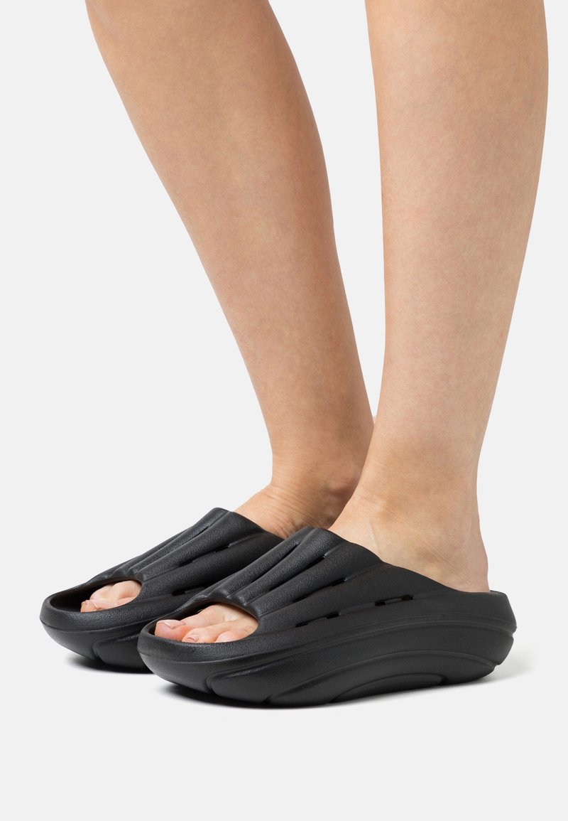 Pieds portant des sandales noires rembourrées, à enfiler, avec des trous de ventilation et des orteils découverts sur fond blanc.