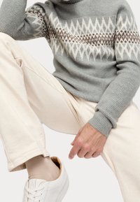 Persoon in een grijs gepatterd sweater, beige broek, beige sokken en witte sneakers, zittend met één knie opgetrokken tegen een witte achtergrond.