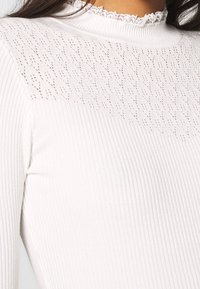 Vit ribbad långärmad topp med ett texturerat, spetsmönstrat ok och en rynkad halslinning som ger en mjuk och ömtålig känsla.