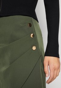Gros plan sur une personne portant une jupe vert olive avec trois boutons dorés sur le côté et un haut noir côtelé à manches longues.
