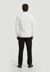 Chemise blanche à manches longues avec une texture lisse, un dos droit, et des poignets à boutons, associée à un pantalon noir et des chaussures.