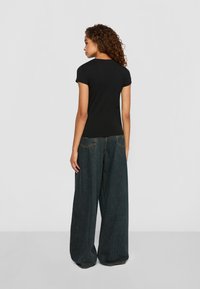Vrouw met krullend haar staat met haar rug naar de camera, gekleed in een getailleerd zwart t-shirt en brede donkere spijkerbroeken tegen een effen achtergrond.