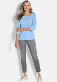 Hellblaues Oberteil mit Dreiviertelärmeln, kombiniert mit grauen Slim-Fit-Hosen und beigen Sneakern. Der Stoff wirkt weich und hat eine glatte Textur.