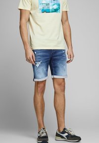 Hombre que lleva una camiseta gráfica de color amarillo claro, pantalones cortos de mezclilla azul desgastados enrollados en el dobladillo y zapatillas en tonos azul marino, blanco y gris.