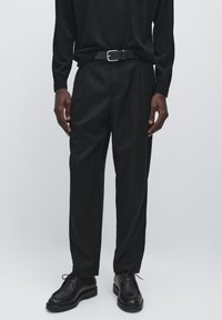 Black long-sleeve shirt paired with black pleated trousers and black leather shoes. Features a black belt with a silver buckle.
