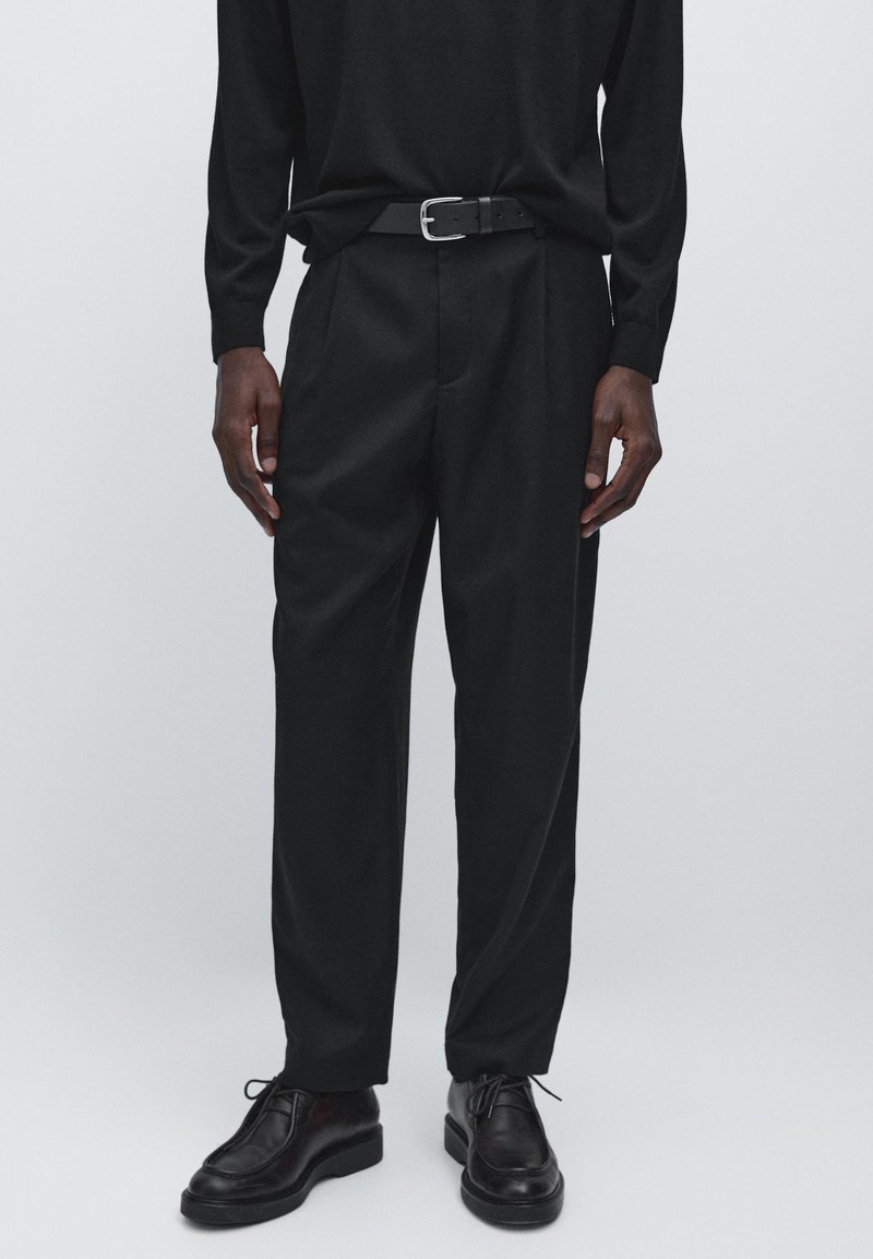 Black long-sleeve shirt paired with black pleated trousers and black leather shoes. Features a black belt with a silver buckle.