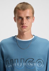 Modrá mikina s vyvýšeným logom "HUGO" v rôznych odtieňoch, okrúhly výstrih, ribbované manžety a plochá textúra. Nosený strieborný náhrdelník.