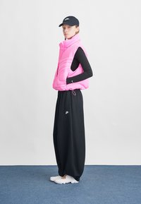 Colete puffer rosa com design acolchoado vertical, top preto de manga longa, calças largas pretas e sapatos brancos, complementados por um boné preto.