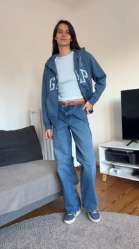 Moteris stovinti patalpoje, vilkinti mėlyną GAP džemperį su kapišonu, šviesiai mėlyną marškinius, plačiomis kojų kelnėmis ir sportbačiais svetainėje su sofa ir televizoriumi.