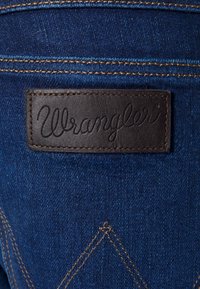 Tamno plavi traper sa istaknutom smešnom kožnom oznakom. Oznaka ima urezanu "Wrangler" oznaku. Kontrastno šivanje duž šavova.