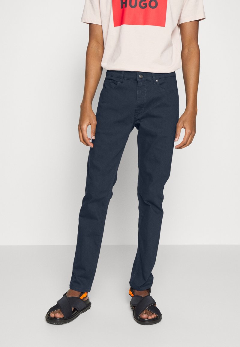 Personne portant un pantalon bleu marine, des sandales noires avec des sangles orange et un t-shirt blanc avec un logo "HUGO" rouge et noir.