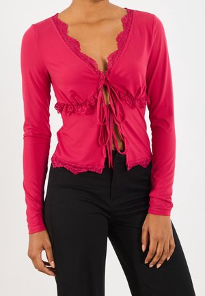 Vrouw draagt een nauwsluitend, lang mouwen roze topje met kanten biesjes en striksluitingen, gecombineerd met een zwarte broek tegen een effen achtergrond.