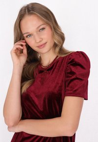 Burgundské sametové šaty s krátkými balónovými rukávy a mašlí na výstřihu. Modelka má vlnité vlasy a nosí korálkový náhrdelník.