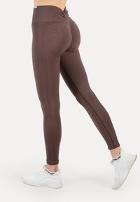 Brązowe leginsy z wysokim stanem, wykonane z teksturowanej tkaniny z bocznymi szwami. Noszone z białymi tenisówkami, podkreślające dopasowany krój i elastyczny materiał.