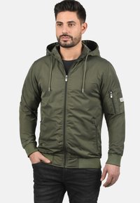 Veste zippée vert olive avec capuche, poignets côtelés et poches latérales ; présente une poche zippée sur le bras pour un rangement supplémentaire.