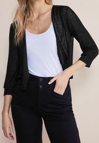 Cárdigan negro, ligero y de abertura frontal con mangas de tres cuartos, usado sobre una camiseta blanca de tirantes, combinado con pantalones negros de talle alto.