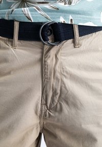 Beige broek met naad in de voorzakken, vergezeld van een zwarte gevlochten riem met een ronde metalen gesp. De stof lijkt lichtgewicht te zijn.