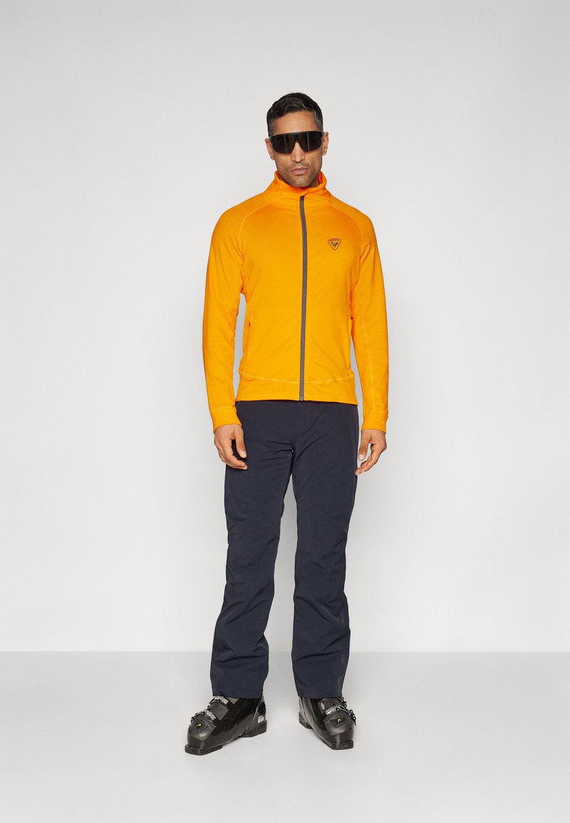 Oranje zip-up jack met een hoge kraag, textielstructuur en logo op de borst, gecombineerd met donkerblauwe broek en zwarte skischoenen.