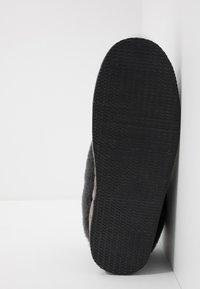 Suela de goma negra con agarre texturizado; parte superior suave de aspecto similar a lana en gris claro con forma redondeada, destacando los detalles de las costuras a lo largo del costado.