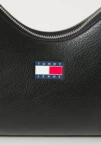 Bolso de mano de cuero negro con superficie texturizada, cremallera plateada y un parche logo rectangular en azul, rojo y blanco que muestra "TOMMY JEANS."
