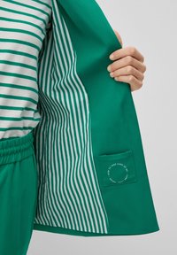 Green jacket with striped interior lining. The outer fabric is smooth, while the inner has thin, vertical white stripes. Features a care label.