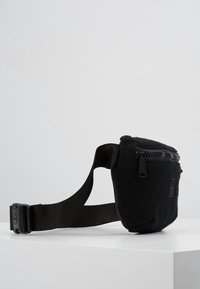 Czarna nerka z polaru z regulowanym paskiem z tkaniny, zamkiem błyskawicznym i płaskim przednim panelem. Kompaktowy design, odpowiedni do przenoszenia niezbędnych rzeczy.