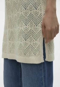 Gebreide top in gebroken wit met een chevronpatroon, voorzien van een afgewerkte halslijn en zijsplitten. Gecombineerd met donkerblauwe denim jeans.