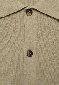 Cardigan lavorato a maglia beige con colletto classico, texture a coste e un bottone circolare scuro sul davanti, che mette in evidenza dettagli sottili.