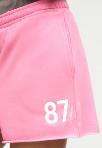 Ροζ βαμβακερό σορτς με ελαστική μέση, πλαϊνή τσέπη και λευκό τύπωμα "87" και υπογραφή στο εμπρόσθιο τελείωμα.
