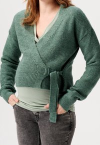 Grön kardi med texturerad stickning, djup v-ringning och knytning i midjan. Bärs över en ljusgrön topp, ihop med mörkgrå byxor.