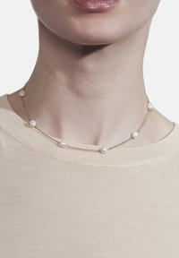Guld halskæde med seks jævnt fordelt hvide perler, båret om halsen, mod en lys beige beklædning.