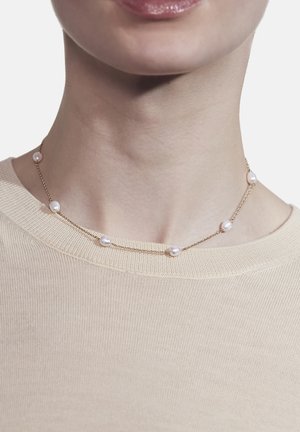 Guld halskæde med seks jævnt fordelt hvide perler, båret om halsen, mod en lys beige beklædning.