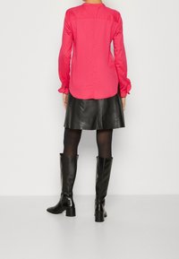 Mujer vestida con una blusa de manga larga rosa, falda de cuero negra, medias negras y botas negras hasta la rodilla, de pie de espaldas con un fondo liso.